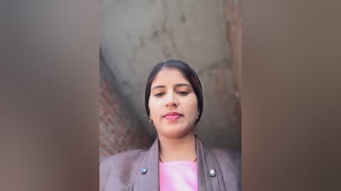 Video of a young South Asian woman with medium brown skin, dark hair, and light makeup, wearing a pink shirt and gray blazer, against a blurred brick wall background.