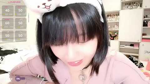 Video of an Asian woman with straight black hair, wearing a pink hoodie with a cartoon dog headband, in a cozy room with shelves and a calendar.
