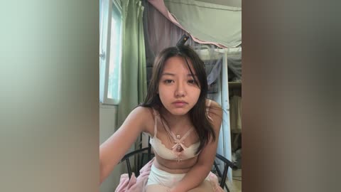 Video of an Asian woman with long black hair, wearing a white lace bra and panties, sitting in a dimly lit room with green curtains and a pink bedspread.