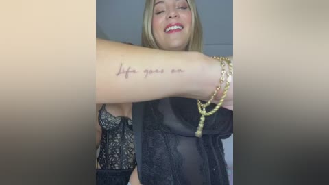 Media: Video of a smiling blonde woman with a tattoo reading \"Life is good\" on her forearm, wearing a sheer black lace dress, and gold chain bracelets.