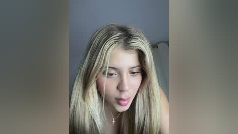 Video of a young, light-skinned woman with long, straight blonde hair, wearing a gold necklace, and making a pouty face. The background is blurred, featuring a white wall and a hint of a door frame.