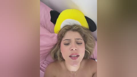 A video shows a topless woman with fair skin, blonde hair, and a yellow Minnie Mouse hat, lying on pink bedding, looking relaxed.