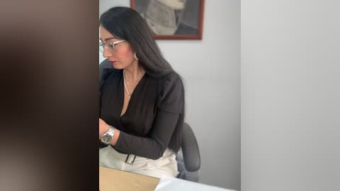 Video of a South Asian woman with long black hair, glasses, and a black blazer, seated at a desk in a minimalist office.