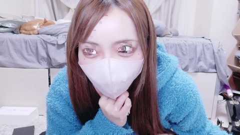 Video of an Asian woman with long, straight brown hair, wearing a white mask, and a blue fleece robe, kneeling on a white and black checkered floor. The background features a gray bed with a brown cat and white curtains.