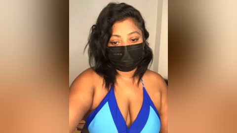 Video of a woman with medium skin tone, wearing a blue halter bikini top, black face mask, and wavy black hair, indoors with blurred background.