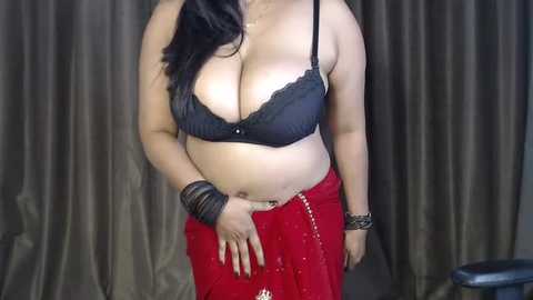 Video of a curvy, light-skinned woman with long dark hair, wearing a black lace bra, red skirt, and black bracelets.