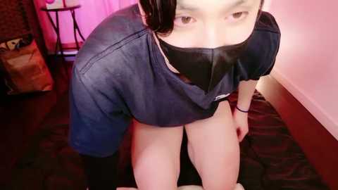 Video of an Asian woman with short black hair, wearing a black mask and dark blue shirt, kneeling on a dark patterned carpet in a dimly lit room with pink curtains and a wooden table.