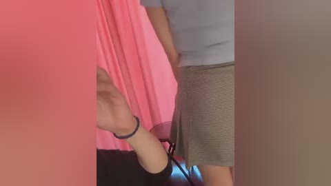 Video of a person's arm in a pink, textured sleeve, wearing a light blue top and grey checkered skirt, standing in front of a pink curtain.