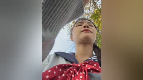 Video of a young Asian woman with light skin, brown hair tied back, wearing a school uniform with a red polka-dotted scarf, looking up at the camera against a blurred background of trees and sky.