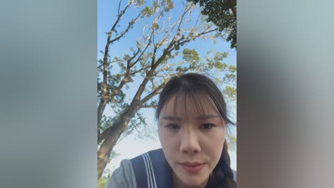 A video of an East Asian woman with straight black hair and bangs, wearing a blue sailor collar school uniform. She stands outdoors under a clear blue sky with a tree's branches and green leaves in the background.