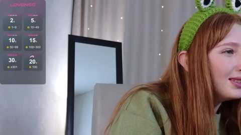 A video of a young woman with long red hair wearing a green knitted headband, smiling. She's indoors, with a weather forecast displayed on a wall and a beige curtain in the background.