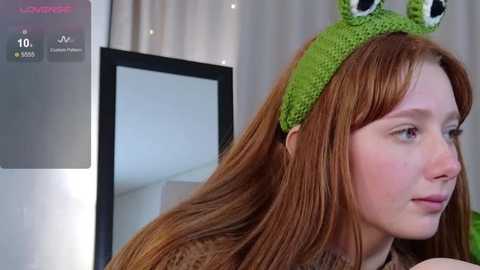 Video of a young red-haired woman wearing a green frog hat, looking contemplative. Background includes a white wall and a partially visible mirror.