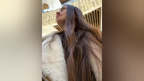 Video of a woman with long, straight brown hair, wearing a white fur coat, looking up at a ceiling with golden chandeliers. The background features a beige and gold color scheme, creating a luxurious, upscale atmosphere.