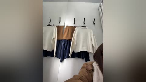 Video of a closet with beige walls, displaying four hanging clothes: two beige sweaters, a brown skirt, and a blue skirt, all on black hangers. A beige blanket is draped on a bed in the foreground.