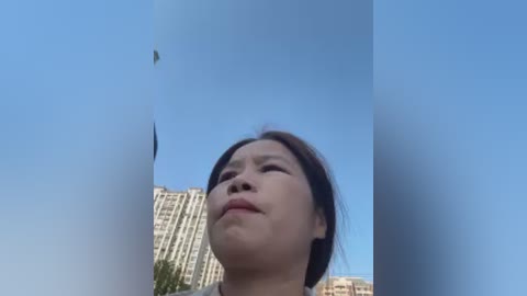 A close-up video of a young Asian woman with straight dark hair, looking upward at a green kite against a clear blue sky. Behind her, modern high-rise buildings are visible.