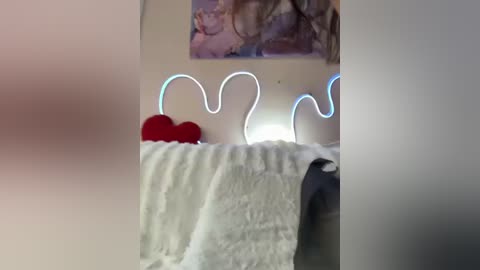 A blurry video of a bed with white sheets, a red heart pillow, and a glowing heart-shaped nightlight. The background includes a soft, blurred image of a painting.