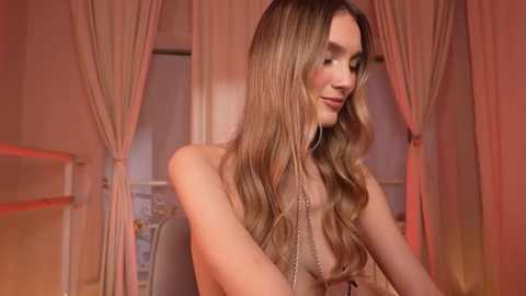 Video of a slender, fair-skinned woman with long, wavy blonde hair, wearing a sheer, beige lace top, seated indoors with light pink curtains in the background, bathed in warm, ambient lighting.