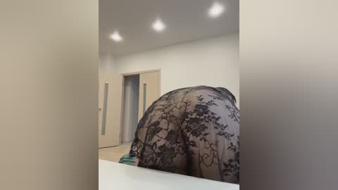 Video of a woman with a large, curvaceous bottom, wearing a black lace dress with floral patterns, bending over a white surface in a minimalist room with beige walls and a door.