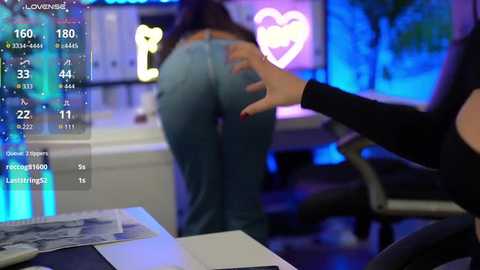 Video of a woman bending over a desk, wearing tight jeans, with a hand on her buttocks, viewed from behind. Virtual reality headset displaying digital health data.