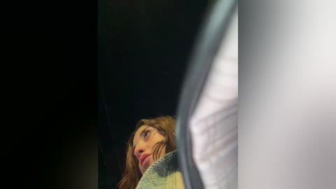 Video of a young woman with long, curly brown hair, wearing a green sweater, looking upward, with a blurred, out-of-focus man's white shirt in the foreground. Dark, dim background.