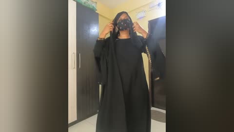 Video of a person with medium skin tone, wearing a black mask, black headscarf, and long black robe, standing in a room with beige walls and dark wooden cabinets.