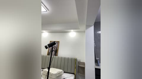 Video of a minimalist, modern bedroom with white walls, a padded headboard, a white bed with neatly folded sheets, and a black microphone stand with a microphone in the foreground.