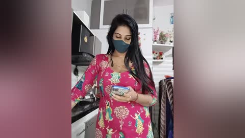 Video of a woman in a bright pink floral dress with long sleeves, wearing a blue face mask and holding a box, in a modern kitchen with white cabinets, appliances, and pink decorations.