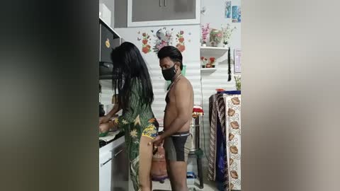 Video of a shirtless, mask-wearing man in black shorts, standing behind a woman in a floral dress, cooking in a kitchen with floral wallpaper and a colorful apron.