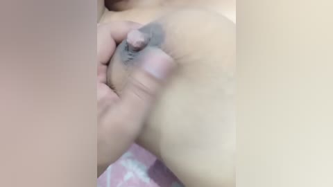 A video of a close-up of a woman's bare breast, with a hand gently squeezing the nipple, which is erect. The background is blurred, showing a pink and white patterned fabric.