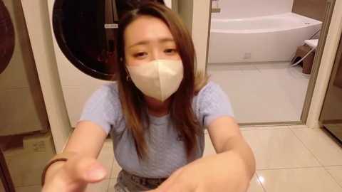Video of a woman in a light blue shirt, beige face mask, and light skin, sitting on a tiled floor in a modern, beige-tiled bathroom with a white bathtub and laundry machine visible.