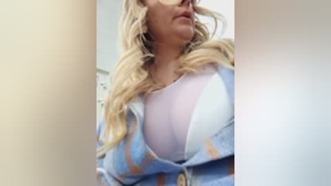 A video shows a blonde woman with long, wavy hair, wearing a white bra under a light blue plaid shirt, partially unbuttoned. The background is a blurred, neutral-toned room.