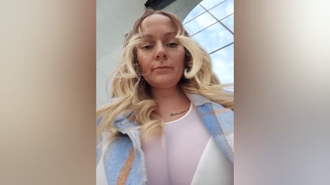 Video of a woman with wavy blonde hair, wearing a pastel plaid shirt over a white top, standing indoors near a large window.