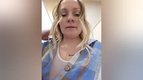 A video of a blonde woman with a fair complexion, wearing a blue and white plaid shirt over a white tank top, standing indoors with blurred background.