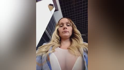 A video of a blonde woman with long hair, wearing a sheer white top that reveals her cleavage, standing in a bathroom with dark tiled walls.