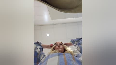 Video of a blonde woman in a plaid shirt, arms raised, standing in a brightly lit room with white walls and a large circular ceiling light.