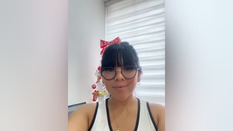 Video of a young Asian woman with black hair and bangs, wearing large-framed glasses, a white tank top, and a red bow, standing in front of a white window with blinds.
