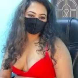 Video of a woman with long, curly dark hair, wearing a red bra and black mask, seated on a black chair, with a blurred background featuring a stained-glass window.