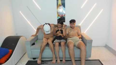 Video of three shirtless men and a woman in a modern living room, lounging on a light-colored sofa; the men are muscular, one in a white hat, the woman in a black top and shorts, all engaged in conversation.