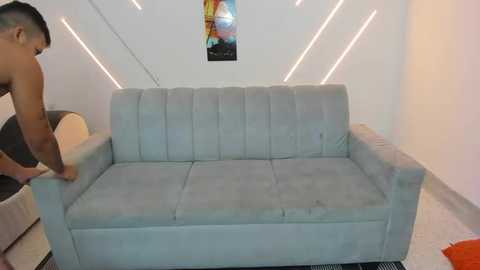 Video of a muscular, shirtless man adjusting a light gray, tufted sofa in a modern, minimalist living room with white walls and recessed lighting.
