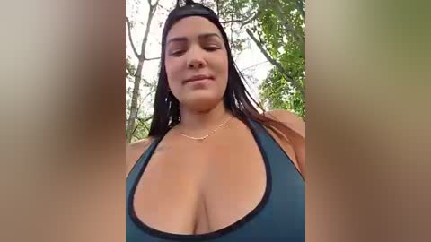 A close-up video of a curvy, light-skinned woman with long dark hair, wearing a black sports bra, standing in a forest with greenery.