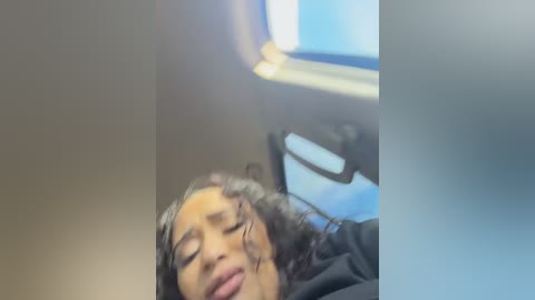 Video of a young woman with curly hair, lying with eyes closed on a black jacket, inside a dimly lit airplane cabin.