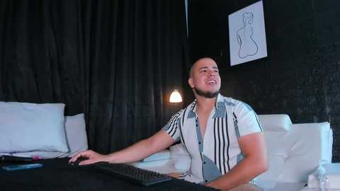 Video of a muscular, smiling man with a beard in a white-striped shirt, typing on a keyboard in a dimly lit, modern room with black curtains, a white bed, and minimalist art on the wall.