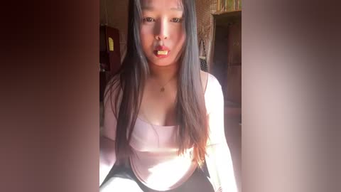 Video of an East Asian woman with long black hair, wearing a light pink top, sitting indoors. She has a light skin tone and is sucking on a lollipop. The background features a dark room with dim lighting.