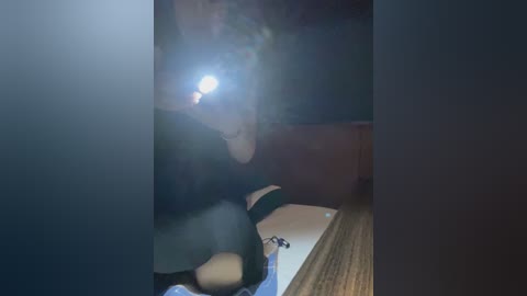 Video of a person with a flash camera in a dimly lit room, partially obscured by shadows. The image is blurry and grainy, with a wooden table and a dark background visible.