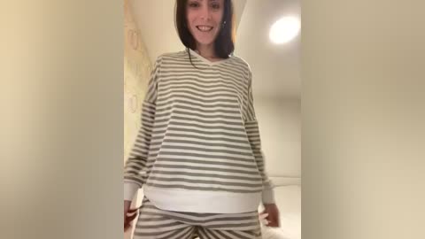 Video of a smiling woman with fair skin and straight dark hair, wearing a loose-fitting, long-sleeved, gray-and-white striped pajama set, standing in a warmly lit, beige-walled hallway.