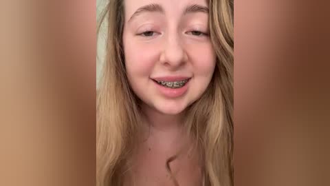 Video of a young, smiling Caucasian woman with long blonde hair, fair skin, and braces, against a blurred, peach-toned background.