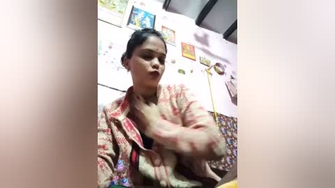 Video of a woman with medium skin tone, dark hair, wearing a patterned beige sweater, sitting in a cluttered room with colorful posters and framed art on the wall.