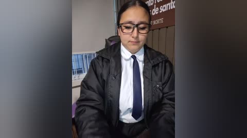 Video of a young woman with dark hair, glasses, and a black jacket over a white shirt and navy tie, sitting indoors with a blurred background.