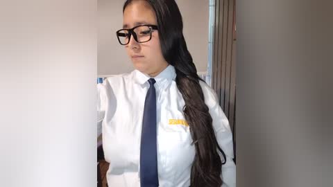 Video of a young woman with long black hair, wearing glasses, a white shirt, and a navy tie, standing against a plain white wall, looking thoughtful.