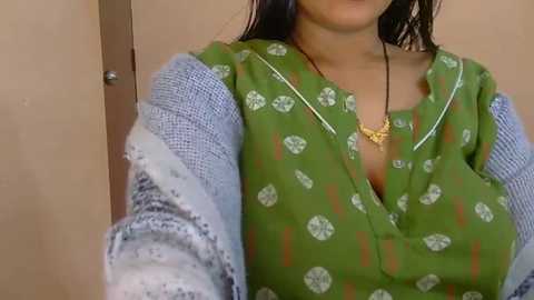 A video of a woman with medium skin tone, wearing a green kurta with white circular patterns, a blue shawl draped over her shoulders, and a yellow necklace. The background is a plain beige wall.